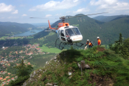 Ein Hubschrauber schwebt über einem Berggipfel. Zwei Menschen stehen auf dem Gipfel. Im Tal ist ein Dorf.