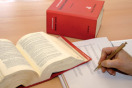 Aufgeschlagenes und geschlossenes rotes Buch sowie Hand mit Stift auf Papieblock auf einer Tischplatte