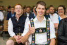 Junger Mann in Tracht mit Hosenträger zeigt mit dem Daumen nach oben. Im Hintergrund sind weitere Meister.