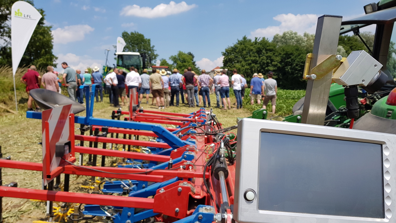 Im Vordergrund steht ein Traktor mit Sähmaschine und Bildschirm. Im Hintergrund stehen viele Personen.