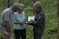 Försterin zeigt einem älteren Ehepaar etwas auf ihrem Laptop im Wald.