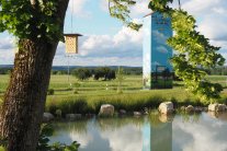 Weiher mit Baum im Vordergrund an dem ein hölzernes Insektenhotel angebracht ist, im Hintergrund ein bemaltes Trafohaus.