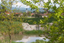 Wasserrückhaltebecken mit Stau- und Ablassbauwerk. Im Hintergrund befindet sich ein Dorf.