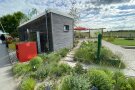 Holz Tinyhouse eingebettet in eine Staudenmischung trägt auf dem Dach freistehend die Worte Zeit für Grün. Es ist sonnig-bewölkt. Ein gepflasterter Weg führt recht neben dem Gebäude zu einem kleinen Platz mit Sonnenschirmen.