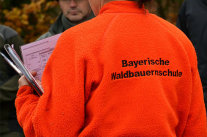 Rückansicht einer Person mit ornger Jacke. Aufschrift: Bayerische Waldbauernschule.