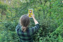 Person hält im Wald ein gelbes rechteckiges Gerät in die Luft.
