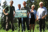 Drei Männer in grüner Uniform und eine Frau in Freizeitkleidung stehen auf einer Lichtung im Wald und halten eine Grafik in Händen