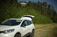 Eine Drohne schwebt über einem Kiefernwald. Daneben steht ein weißes Geländefahrzeug.