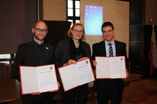 Zwei gut gekleidete Herren und eine Dame präsentieren sich mit Urkundenmappen in einem Festsaal dem Fotografen.