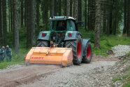 Traktor mit Anbaugerät zerkleinert Steine auf einem Forstweg.