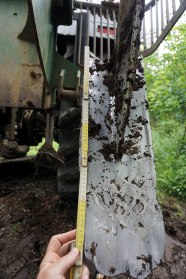 Ein Metallteil, das wie ein Spaten wirkt, hängt an einem Traktor; zum Größenvergleich wird ein Meterstab daneben gehalten; das Metallblatt ist groß