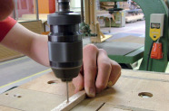 Der Arm eines jungen Menschen hält ein Holzstück, in welches mit einer Maschine ein Loch gebohrt wird. 