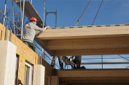 Bauarbeiter lenkt vom Kran bewegtes großes Holzteil auf einer Baustelle eines Fertigteilhauses aus Holzelementen.