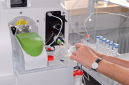 Zwei Hände hantieren an einem Gerät im Labor. Daneben steht eine Palette mit Reagenzgläsern.