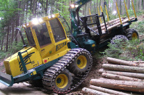 : A forwarder with eight wheels and bogie tracks is driving backwards from a skid trail on a fairly steep slope towards a forest road.