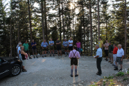 Menschen stehen im Halbkreis um einen Vortragenden auf einem Schotterplatz im Wald