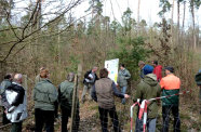 Ein Mann hält mit Hilfe einer Schautafel vor ein paar Leuten einen Vortrag im Wald.