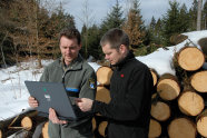 Zwei Männer stehen draußen im Schnee vor einem Holzpolter und gucken in ein Laptop, das der Mann mit Forstuniforn in Händen hält