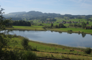 Langgezogener kleiner Seee in hügeliger Landschaft mit Grünland.