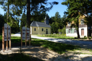 Kirchlein und altes Haus im Wald. Im Vordergrund steht eine Infotafel.