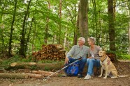 Älteres Paar mit großem Hund sitzt auf einem Waldweg auf einem Eichenstamm..