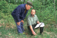 Das Bild zeigt zwei Männer, die auf einer Lichtung im Wald stehen. Einer der Männer ist blau gekleidet und steht in einer gebeugten Haltung, während er dem anderen knieenden Mann zuhört.
