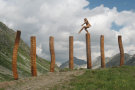 Sieben Holzstelen im alpinen Kontext, darauf geht ein Mann - Skultpur aus Holz