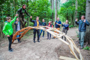 Menschen stehen im Wald und schauen einer Frau zu, die über eine selbst gebastelte Brücke geht, während ein Mann ihr die Hand zur Stabilisierung reicht