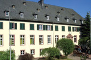 Das Bild zeigt ein weißes, vierstöckiges Gebäude mit grünen Fensterläden und grauem Dach. Vor dem Gebäude stehen vier kleine Bäume. Im Hintergrund ist der Himmel blau.