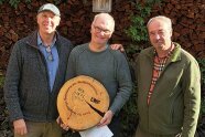 Drei Männer stehen für ein Gruppenbild vor aufgeschichtetem Holz.