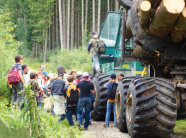 Das Bild zeigt eine grüne Holzrückemaschine auf einem Forstweg. Auf der linken Seite der Maschine stehen vielen Kinder. Im Hintergrund sieht man Wald.