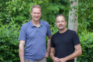Zwei Männer in Hemdsärmeln stehen nebeneinander vor Birkenwald