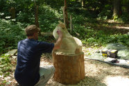 Ein Mann mit blauem Tshirt und Käppi sitzt auf dem Boden und arbeitet an einer Skulptur aus Holz