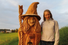 Eine junge Frau steht neben dem selben Holzklotz, nur dass er nun fertig bearbeitet ist und eine Hexe aus dem Märchenbuch zeigt