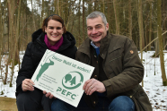 Ein Mann und eine Frau knien im Winter vor einem jungen Laubwald und halten ein Schild in die Kamera.