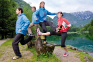 Vier Personen beim Freiluftfitnestraining an einer Ruhebank an einem See. Im Hintergrund sind Berge zu sehen.