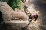 Frau bearbeitet mit Stechbeitel und Holzhammer einen Holzstamm. Es entsteht eine Skulptur.