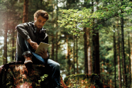 Ein junger Mann (hipster) sitzt auf einem Baumstumpf und bedient ein Tablet in einem Mischbestand
