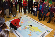 Eine Gruppe von Personen steht um eine große auf dem Boden ausgebreitete Weltkarte herum. Eine Frau kniet darauf.