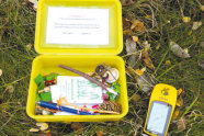 Gelbe Plastikdose mit Stiften und weiteren Kleinteilen. Daneben liegt ein GPS-Gerät.