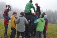 Eine Gruppe Jugendlicher auf einer Wiese am Waldrand. Einige stehen auf einem Stück Pappe, der Rest darum herum. Drei von ihnen sitzen bei anderen auf der Schulter.