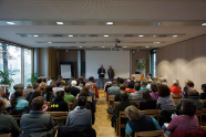 Ein großer Saal in dem zwei Menschen vorne bei einer Präsentationsfläche stehen, und etwa 30 andere Menschen auf Stühlen sitzen und nach vorne blicken