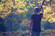 Ein hipper Dude mit Manbun steht in Rückenansicht am Waldrand, die Sonne scheint in die Kamera. Der Typ hat den rechten Arm erhoben und hält sich damit an einem Eichenast fest. An diesem Arm trägt er ein echt trendy Armband.