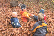 Kinder sitzen in tiefem Herbstlaub. Sie befüllen einen Korb, zwei weitere Körbe liegen etwas abseits.