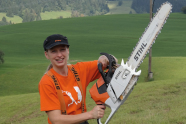 Eine junge Frau im orangenen Shirt steht auf einer Wiese und hält eine Motorsäge, fast so groß wie sie selbst, in die Luft