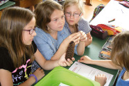 Vier Mädchen sitzen an einer Schulbank, auf der Hefte, Mäppchen und ein Zapfen liegen.