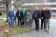 Sieben Männer stehen um einen Stein mit dem Portrait eines anderen Mannes