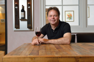 Mann sitzt an einem Tisch, hält ein auf dem Tisch stehendes halb volles Glas Rotwein mit der rechten Hand.